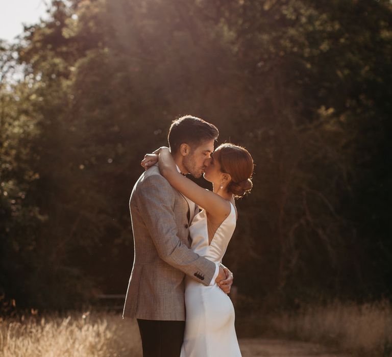 Bride in fitted silk wedding dress embraces her groom in grey suit jacket 