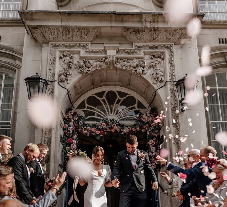 Bride in satin v neck wedding dress with puff sleeves and front slit and groom in black tux walking through confetti at Somerset House