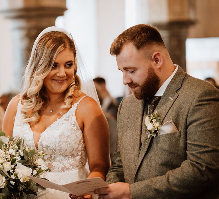 The groom looks at the order of service booklet while the bride smiles at him adoringly during their church ceremony 