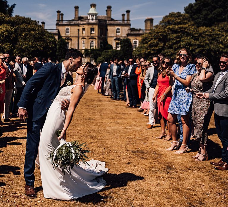 The groom in a navy suit and pink tie leans the bride as he kisses her at the end of their confetti exit 