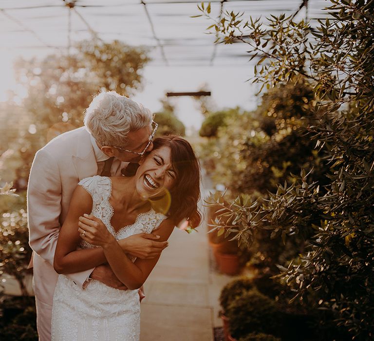 Groom hugs Bride from behind and kisses her on forehead while she laughs with greenery in the background