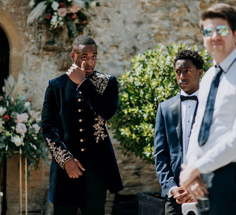 Groom in midnight blue and gold Indian wedding outfit wipes tear away from eye in emotional reaction to bride