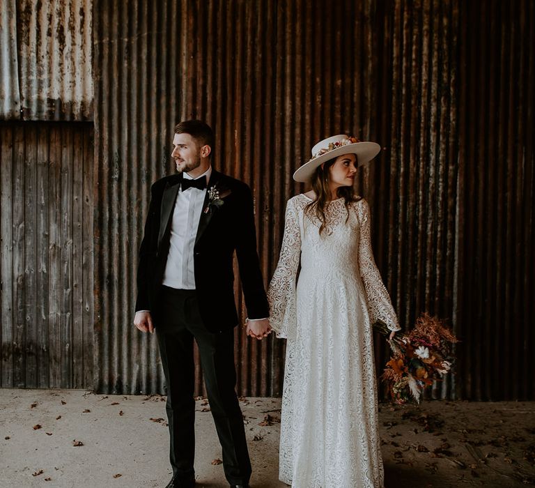 Groom in velvet tuxedo with the bride in a laser cut lace wedding dress with bell sleeves and a white hat carrying a colourful bouquet 