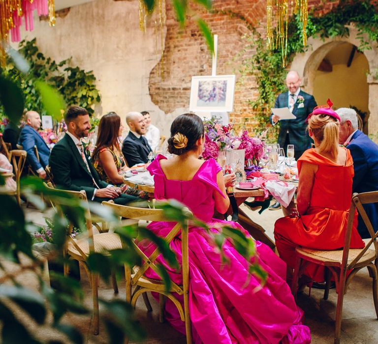 Bride in off the shoulder front ruching pink wedding dress and other wedding guests listening to groom in dark forest green tartan suit with patterned tie, white pocket square and pink and green boutonniere giving wedding speech