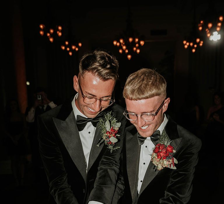 Grooms in matching black velvet suits with bowties and red rose and foliage boutonnieres cutting into their five tier wedding cheese tower with grapes, figs, red roses and foliage decor