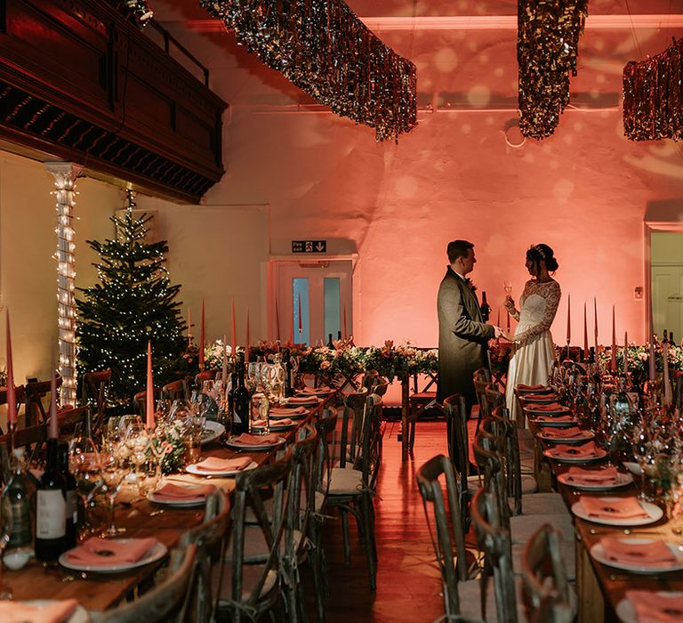 Bride and groom sharing a glass of wine in the reception room of London wedding venue with gold wedding streamers, projections, orange tapered candles, orange napkins, orange rose and foliage tablerunners and fairy lights