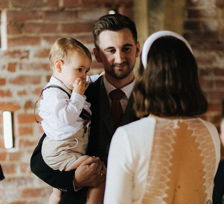 Groom holding the toddler smiles at the bride wearing an open back wedding dress with lace detailing for the civil wedding ceremony at Grangefields wedding venue