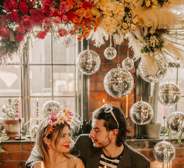 Bride and groom gazing at each other sitting at colourful wedding tablescape with pink metallic streamers, hanging disco ball decorations, viva magenta, fuchsia and hot pink tapered candles and suspended pink and yellow rose flowers with foliage 