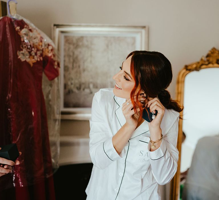 Bride wearing white personalised pyjamas puts on her earrings on the morning of her wedding day