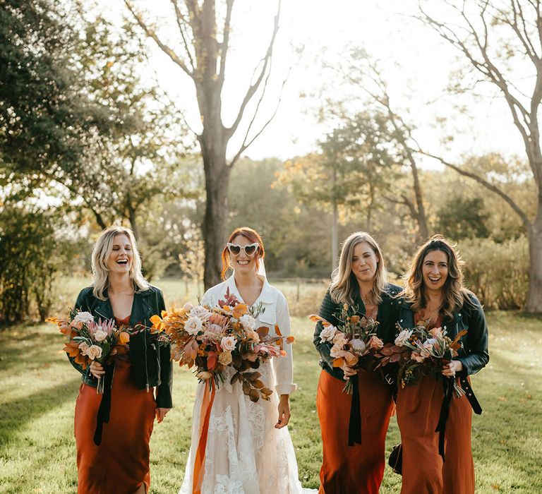 Bride walks alongside her bridesmaids in burnt orange satin bridesmaid dresses and black leather jackets holding orange floral bouquets