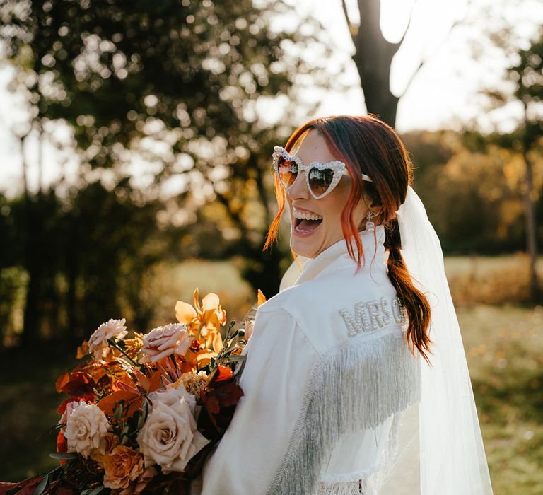 Bride wears heart shaped sunglasses and white custom design denim jacket with glittering tassels and name to the back