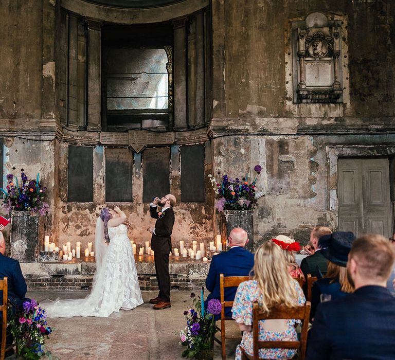 Groom in mocha brown textured suit with deep green tie and pink and purple boutonniere and bride in dainty leaf 3d appliqué sheer bridal gown and cathedral length veil doing shots at the alter as part of their bespoke wedding ceremony 