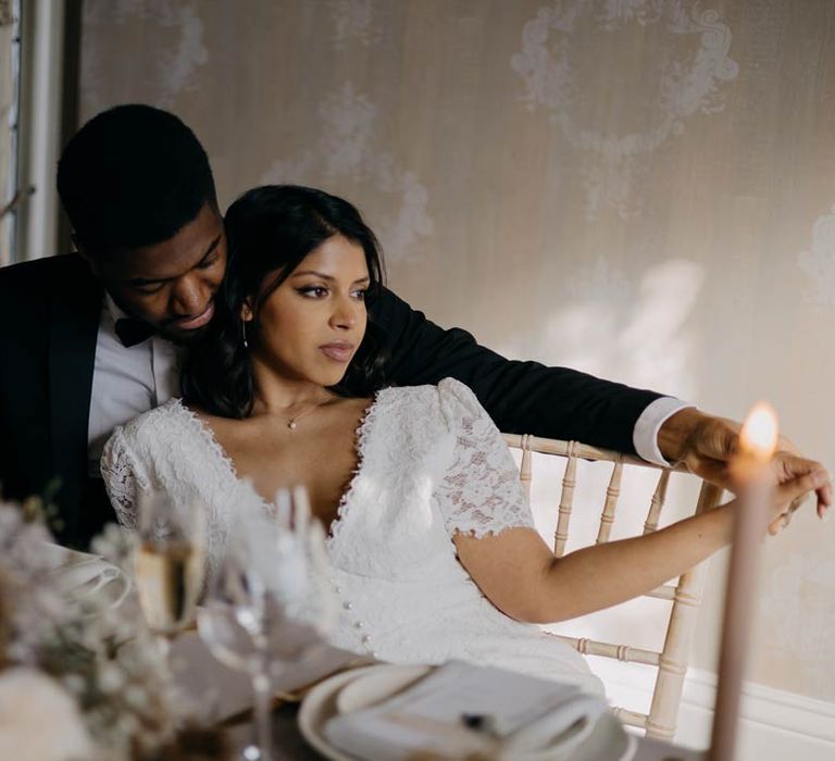 Bride in v neck short sleeve wedding dress with corset-style button detailing sitting at minimalist luxe wedding tablescape sat with groom in classic black tuxedo, black bowtie and white pocket square