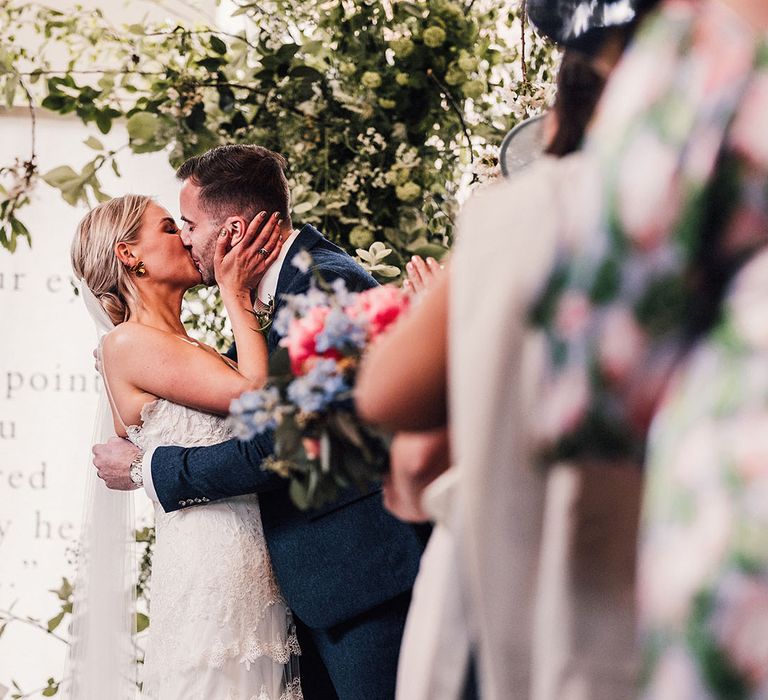 The bride in a lace wedding dress and groom in a blue suit share their first kiss as a married couple 