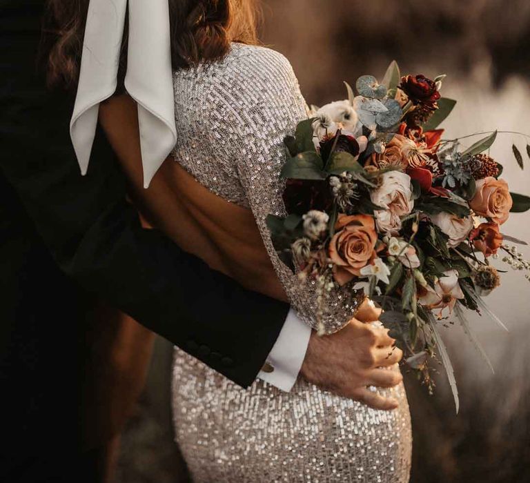 Bride in silver sparkly wedding dress with open back detail and large white satin bow hair bridal hair accessory with groom in classic black tuxedo looking out at the lake holding bouquet with garden roses, carnations, peonies, eucalyptus, baby's-breath and dried flowers tied with mustard velvet ribbon 