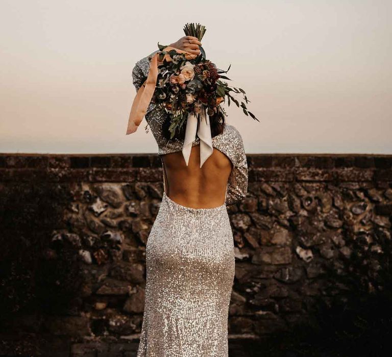 Bride in silver sparkly wedding dress with open back detail holding bridal bouquet behind her head 