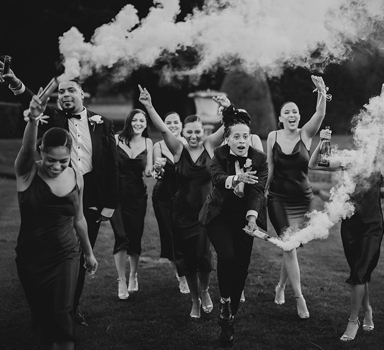 Wedding party in strappy satin midi length black bridesmaid dresses and classic black tuxedos with white rose boutonnieres doing smoke bomb wedding photos at Grittleton House wedding venue 