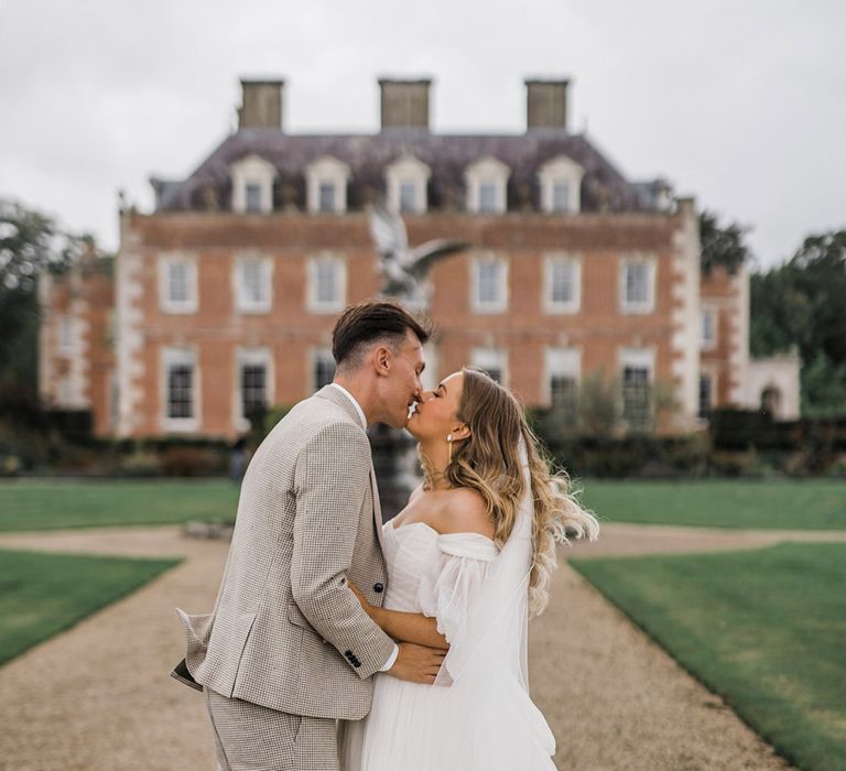 Groom in houndstooth suit kissing the bride in a princess wedding dress on the drive to St Giles House 