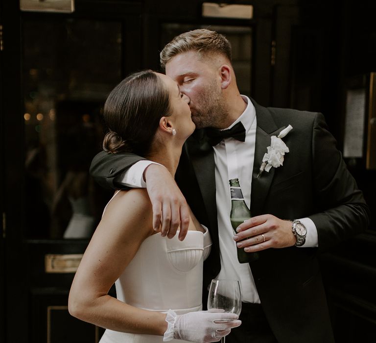 Bride in satin wedding dress with tulle bridal gloves kisses the groom in black tux holding a bottle of beer 