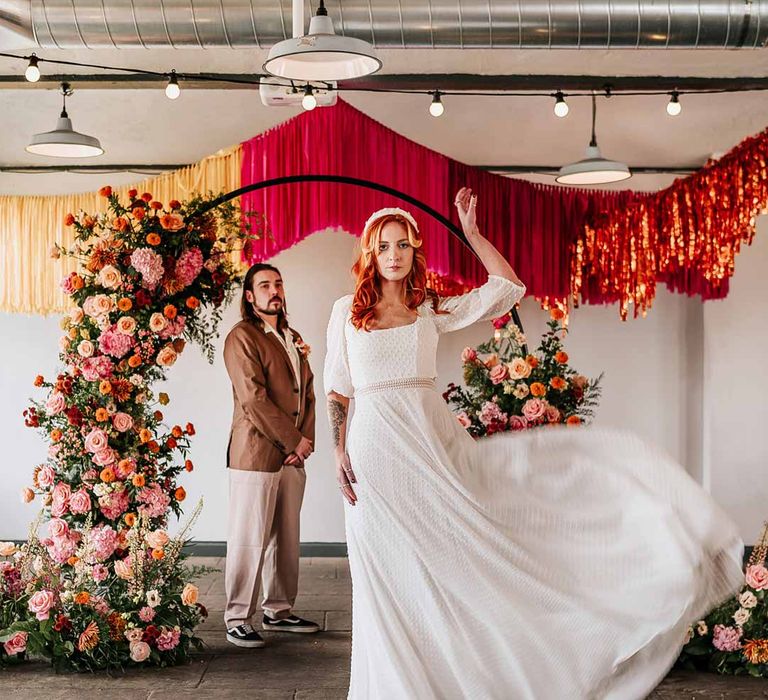 Bride in three quarter length puff sheer sleeve wedding dress with delicate polka dot overlay and square neck and white 3D applique polka dot headband with groom watching on wearing brown blazer, white shirt, grey suit trousers and vans shoes standing underneath circular floral arch with vibrant flowers and foliage attached and pink, red, yellow and metallic wedding streamers behind