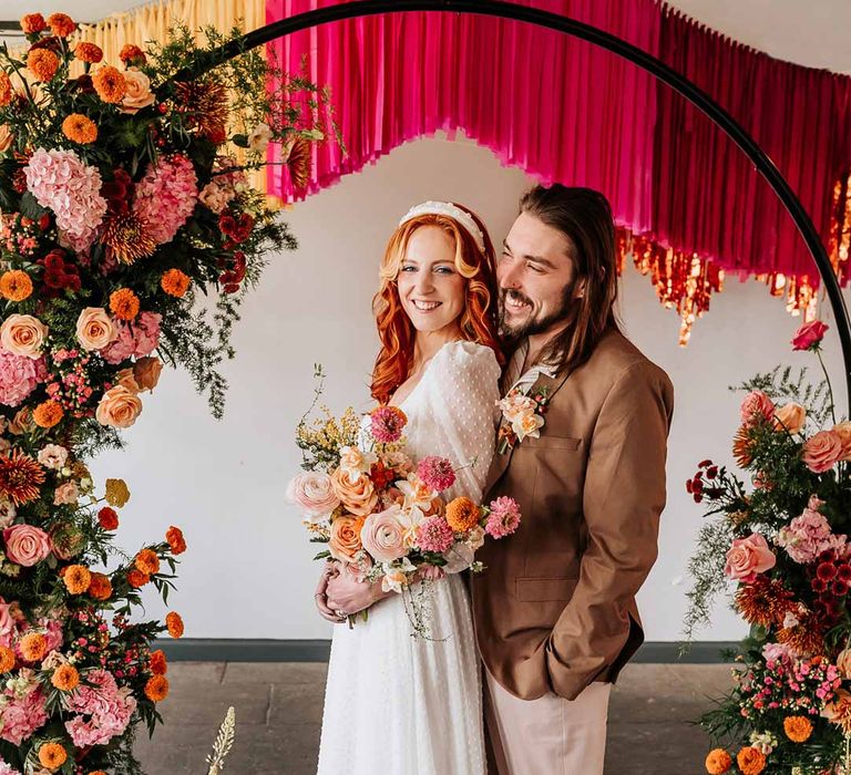 Bride in three quarter length puff sheer sleeve wedding dress with delicate polka dot overlay and square neck and white 3D applique polka dot headband posing with groom in brown blazer, white shirt, grey suit trousers and vans shoes underneath circular floral arch with vibrant flowers and foliage attached and pink, red, yellow and metallic wedding streamers behind at Northern Monk Refectory Leeds