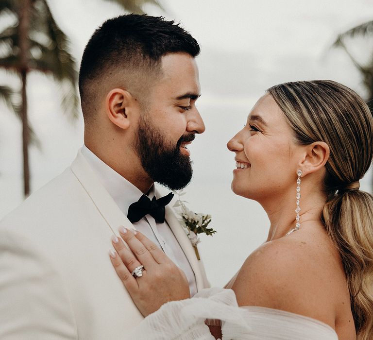 bearded groom in a white dinner jacket and bride in a strapless long sleeve tulle wedding dress with long wavy ponytail hairstyle embracing her husband 