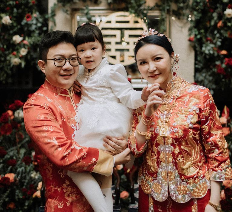 The bride and groom pose with their daughter who wears a long sleeve white dress with pearl necklace accessory 