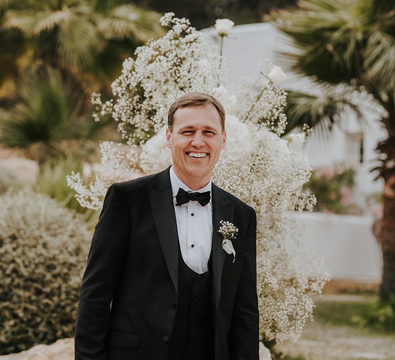 groom in a black tuxedo with horseshoe waistcoat and bow tie smiling at the altar