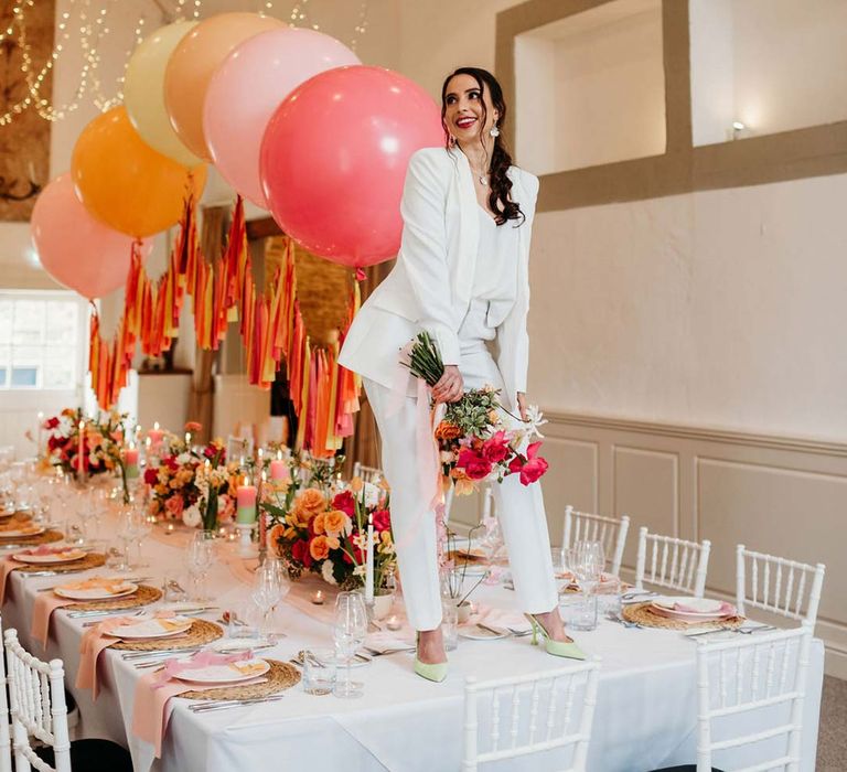 Bride in white bridal suit with silver bridal jewellery with closed toe bright green wedding heels standing on wedding tablescape at Merriscourt wedding venue with wedding tablescape with white wedding tablecloth, burlap place mats, tapered candles, pink and orange wedding stationery, and floral wedding table centrepieces with pink and orange garden roses, orange floribunda, yellow ranunculus flowers, foliage and Iceland poppies with red, pink orange and yellow balloons, wedding streamers and fairy lights at Merriscourt wedding venue