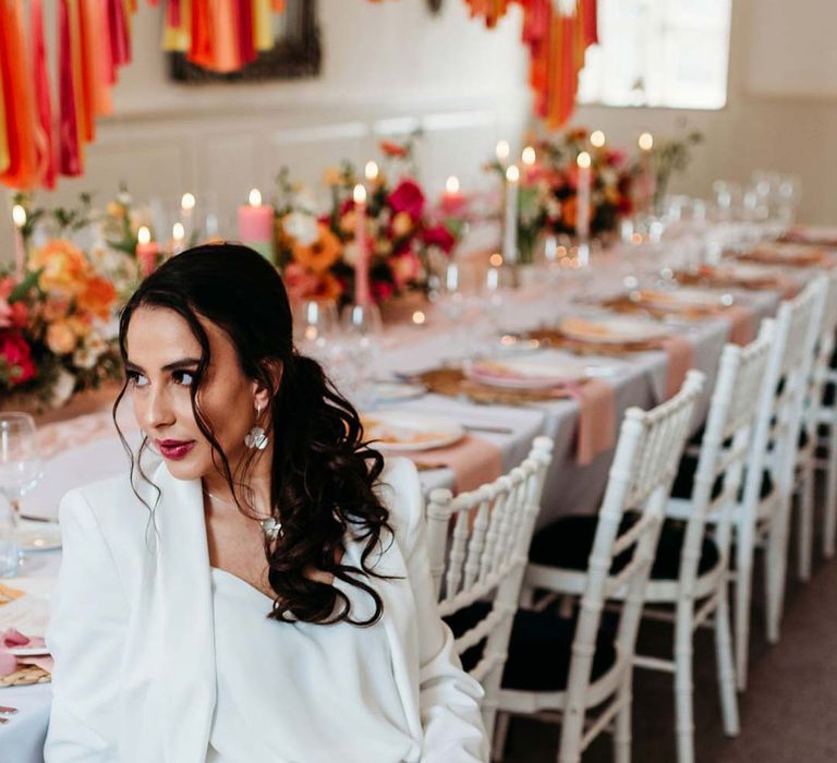 Bride in white bridal suit with silver bridal jewellery sitting in by wedding tablescape with white wedding tablecloth, burlap place mats, tapered candles, pink and orange wedding stationery, and floral wedding table centrepieces with pink and orange garden roses, orange floribunda, yellow ranunculus flowers, foliage and Iceland poppies at Merriscourt wedding venue