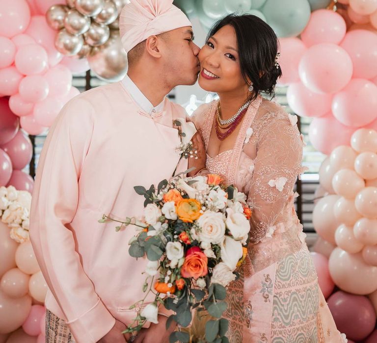 Bride and groom embracing next to large wedding balloon arch in gold, pink, red and baby blue