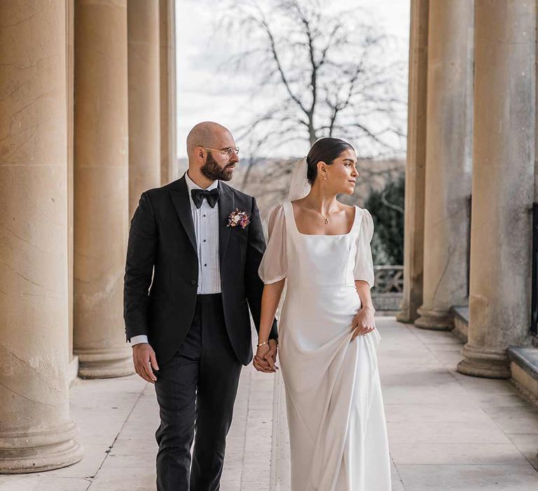 Groom in classic black tuxedo with bowtie and wildflower boutonniere walking with bride in short sheer puff sleeve wedding dress with square neck and wrist loop with sheer pearl embellished bridal veil at Buxted Park