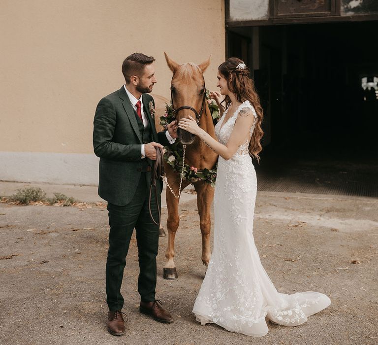 bride in an appliqué wedding dress and groom in a dark green suit with their pet horse in a flower collar at tenuta il cigno, riding school wedding