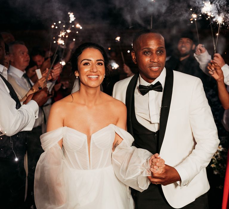 Bride in white tulle wedding dress walks through sparkler exit with groom in white and black tuxedo