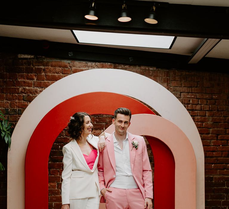 Red, pink and white arches and disco ball photo backdrop with the bride and groom in pink and white suits that match the colour palette 