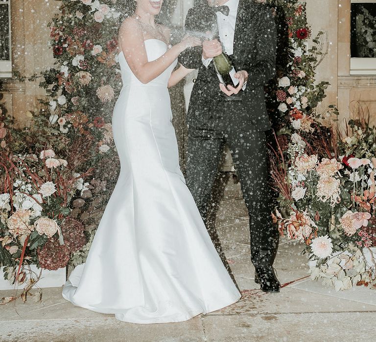 Bride in mermaid wedding dress with groom in black tuxedo popping champagne with flower columns 