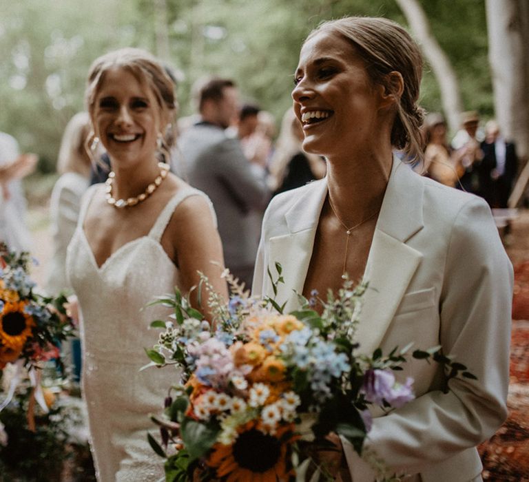 Two brides carrying pastel wedding bouquets exit their ceremony to confetti 