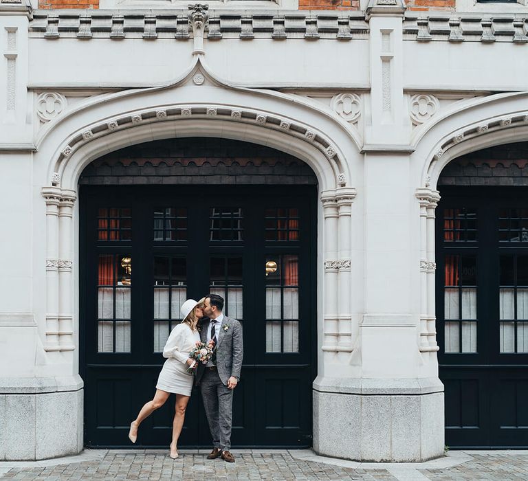 Bride leaning on one leg wearing a short wedding dress to kiss her new husband at London wedding