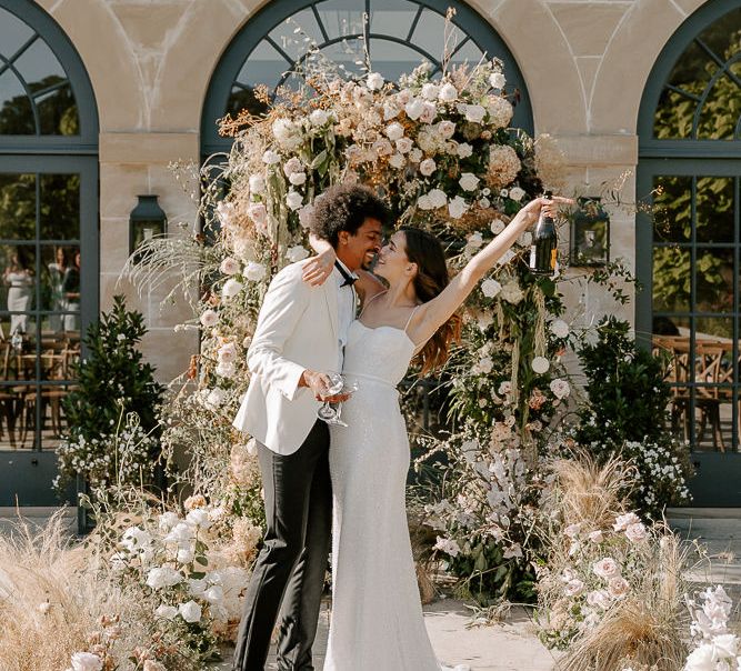 Bride in a sparkly wedding dress with thin straps celebrating with her groom with champagne in front of the flower arch 