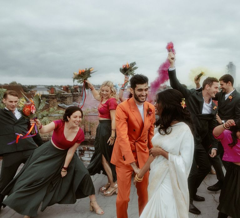 Fun wedding party portrait on the wedding venues roof with bridesmaids in pink and green lenghas, groomsmen in teal suits, groom in an orange suit and the bride in a white sari