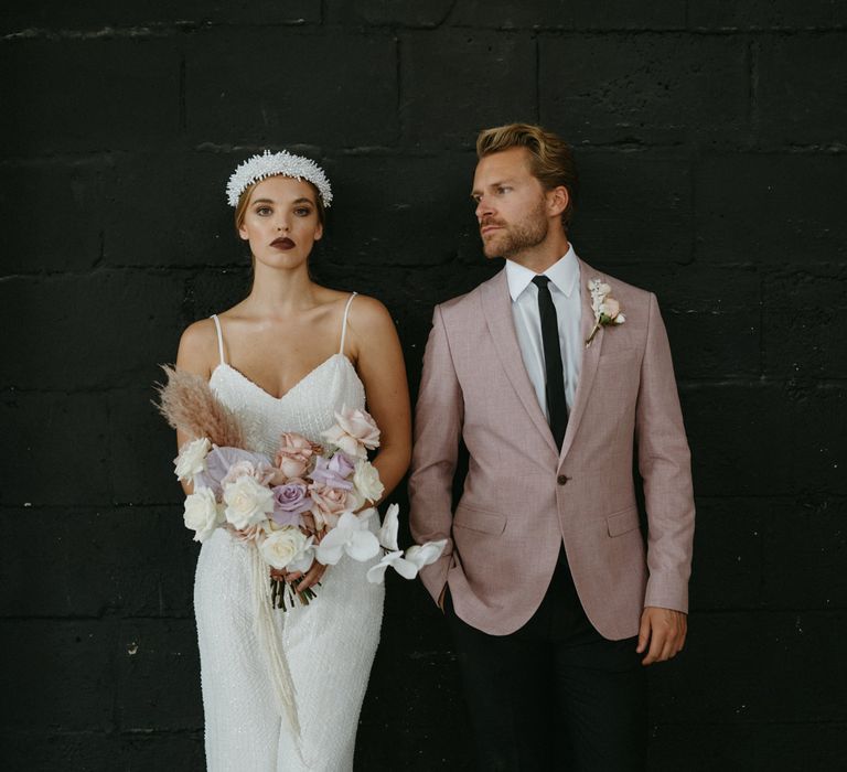 Stylish bride in a white sequin jumpsuit holding a pastel bouquet and groom in a pink blazer standing against a black wall 