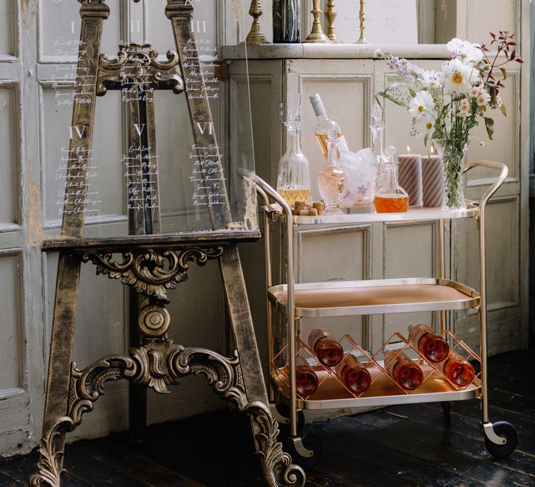 A Perspex table plan resets on an ornate easel. It stands next to a gold bar cart that has glass decanters on top. Photography by Rebecca Goddard.