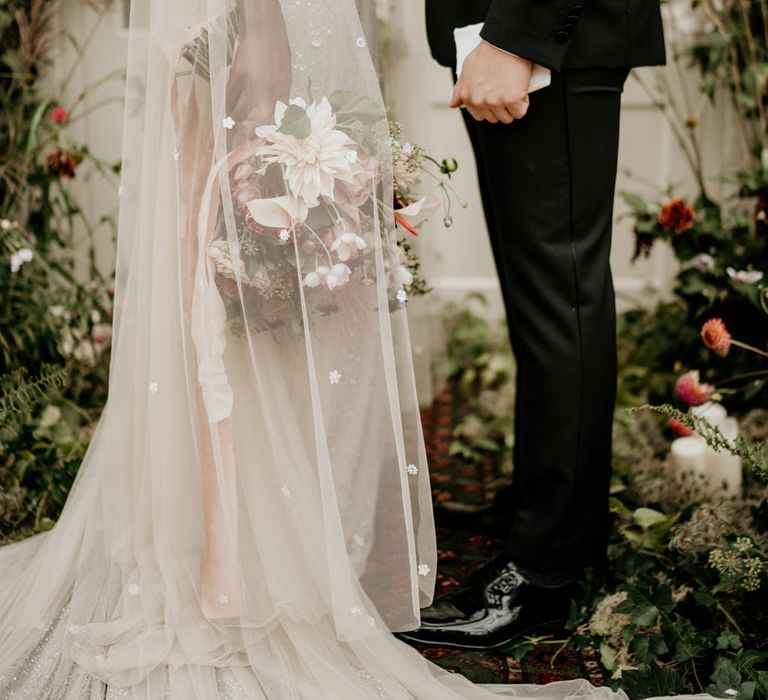 Bride in a Julita Bridal London wedding dress with embellished detail and cape standing at the altar with her groom in a black tuxedo 