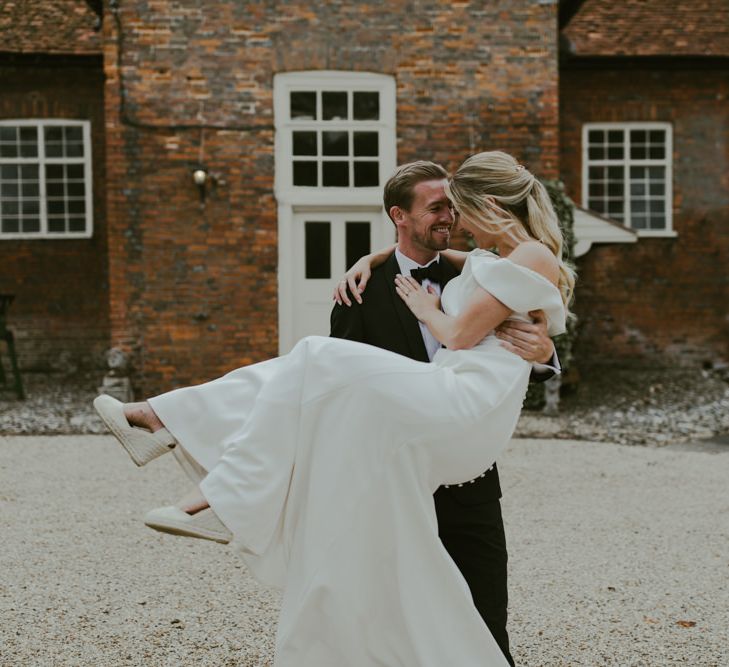 Bride & groom laugh together as he carries her through barn grounds