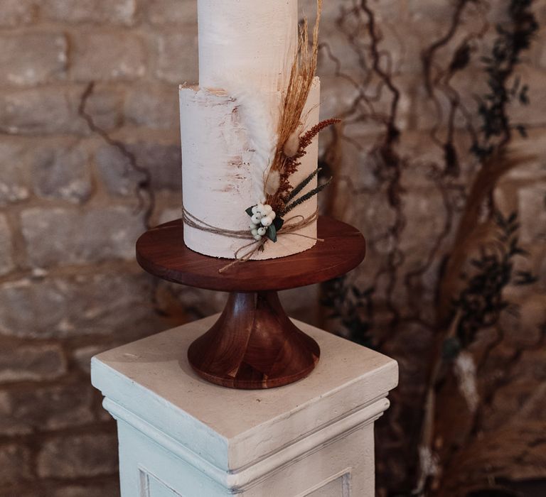 Tall two-tier wedding cake on a wooden cake stand with dried grass tied around the cake for decoration