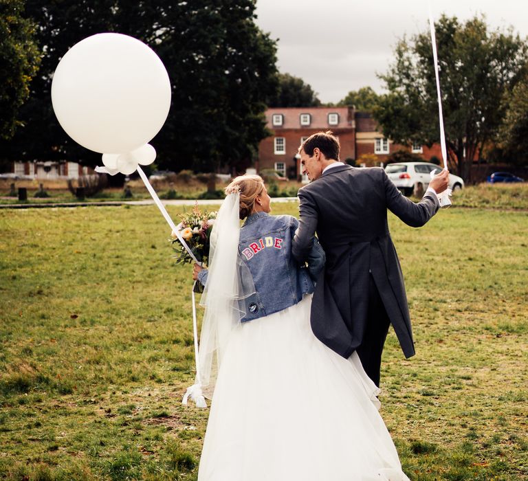 Bride walks with groom whilst she wears personalised denim jacket and they both hold white balloons