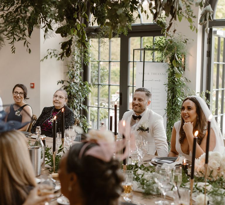 Groom in white tuxedo jacket, bride in Made With Love wedding dress and veil and guests sit at table listening to speeches in the Fig House decorated with greenery