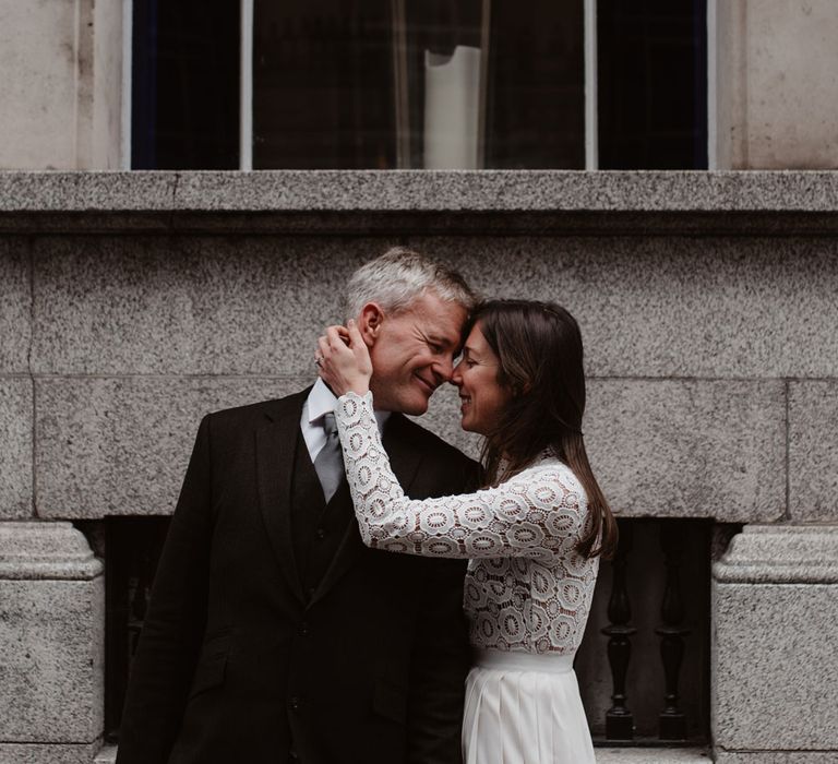 Bride in long sleeved lace top Self Portrait wedding dress holds head of groom in brown woollen blazer and grey trousers outside Bristol Registry Office