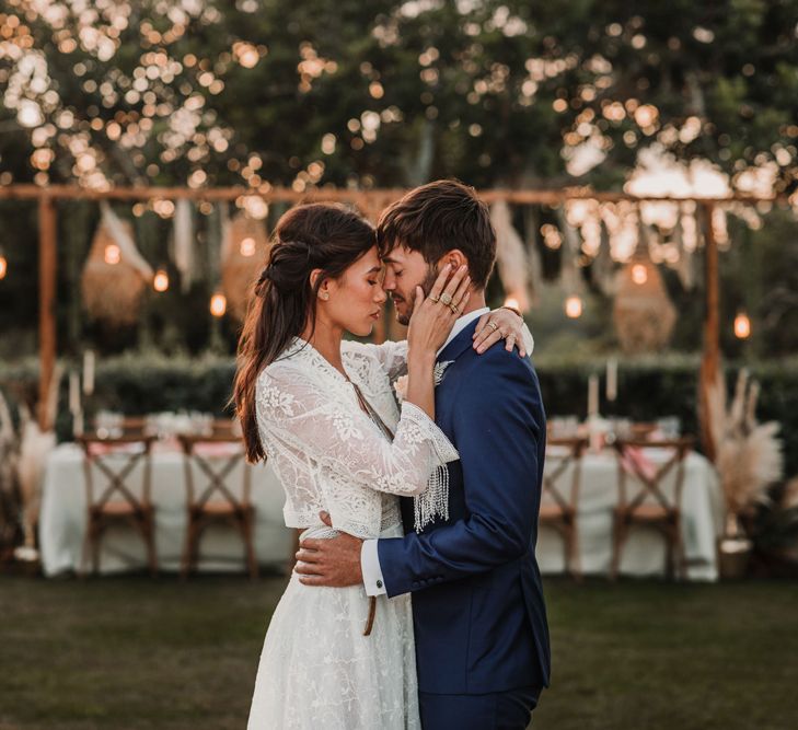 Boho bride in a lace dress with matching jacket embracing her husband in a navy suit at the outdoor wedding reception 