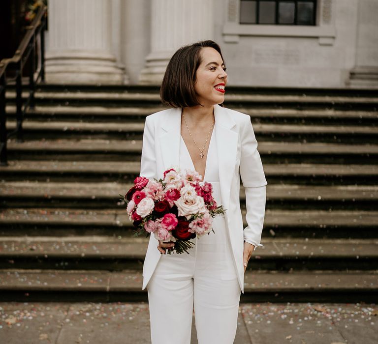 Bride wears white jumpsuit whilst holding pink and red floral bouquet